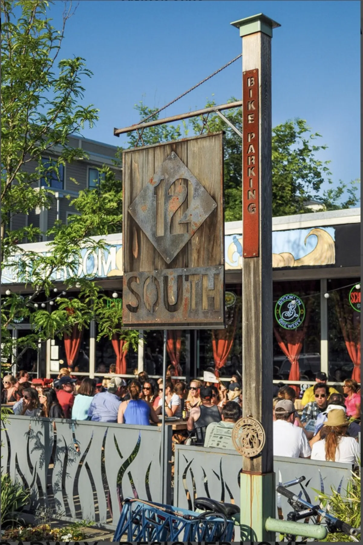 A vibrant exterior scene in the 12 South District, featuring the iconic 12 South sign, a lively crowd of people, and visible bike parking. The setting is bustling with activity, with locals and visitors enjoying outdoor dining and socializing along the popular neighborhood street.
