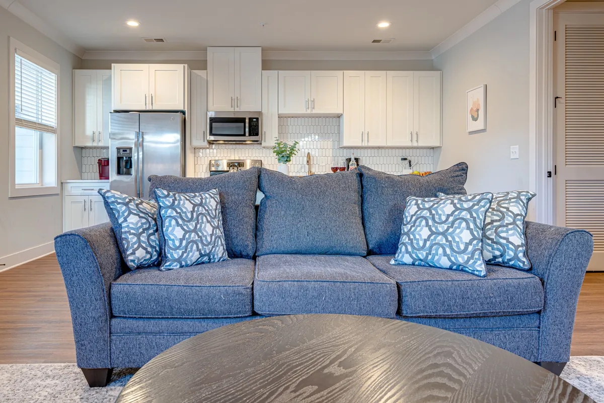 A cozy living room with a plush sofa and pillows, a coffee table, and a view toward the kitchen area. The space is warm and inviting, designed for both relaxing and socializing, with the kitchen easily accessible for entertaining or preparing meals.