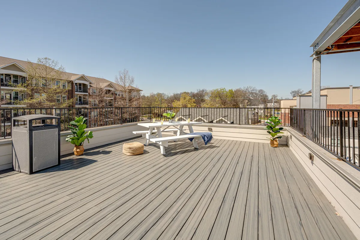 A rooftop deck with a picnic table, planters adding greenery, and deck flooring. The setting is casual yet inviting, perfect for social gatherings, dining, or relaxing. The presence of a trash can suggests a well-maintained communal area. The light hints at either early evening or late afternoon, supporting sunset moments.