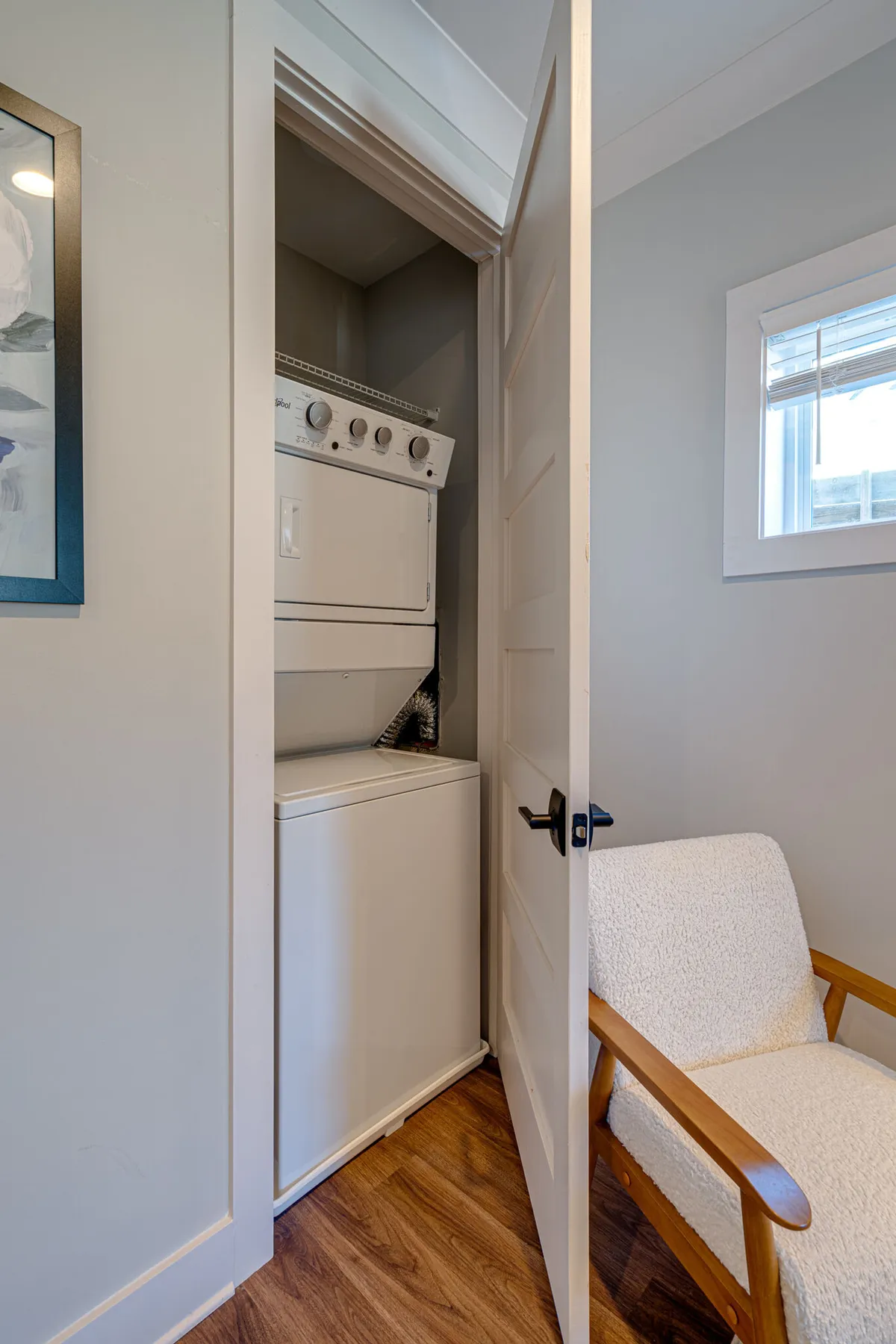 A practical laundry area with a stacked washer and dryer, adjacent to a comfortable chair and framed artwork. The space is functional yet inviting, emphasizing ease of daily routines.