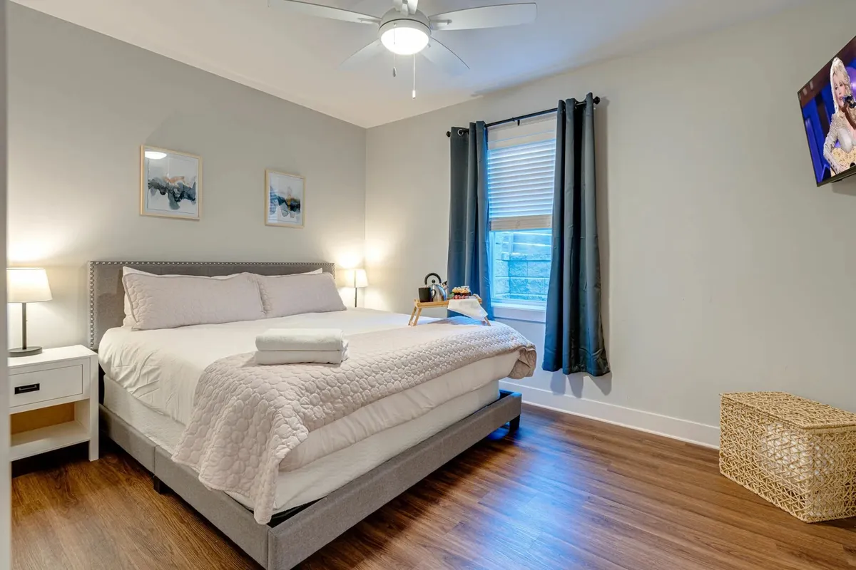A cozy bedroom featuring a king-size bed adorned with plush bedding and decorative pillows, a nightstand with a lamp, a large window allowing natural light to pour in, a wall-mounted television, and tasteful artwork above the bed. The overall palette and decor create a calming, serene ambiance ideal for relaxation.