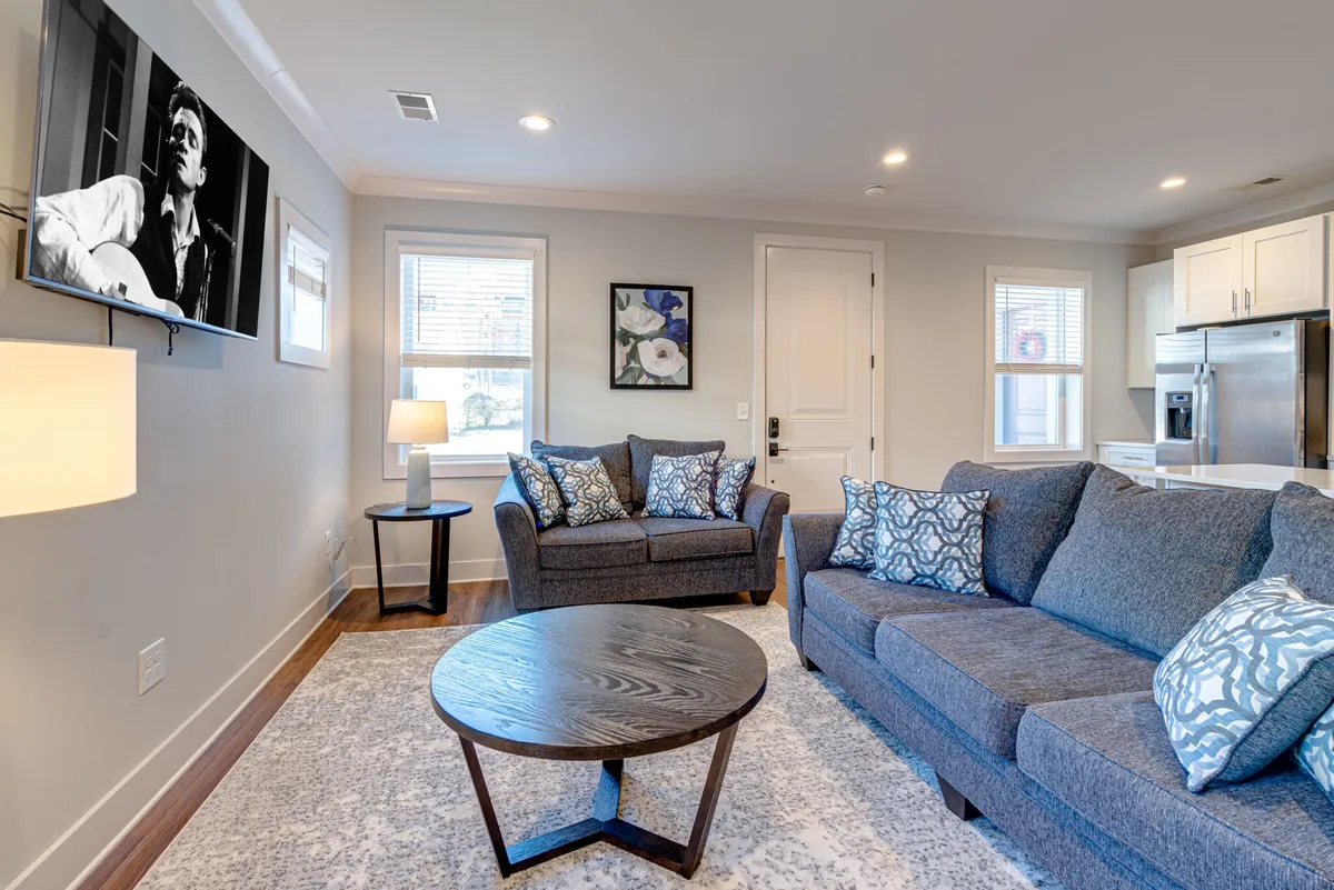 The image showcases a cozy, stylish living area with a plush sofa adorned with decorative pillows, a modern coffee table, vibrant wall art, a soft-glowing lamp, and a TV featuring a musician—inviting guests to unwind and enjoy quality time together.