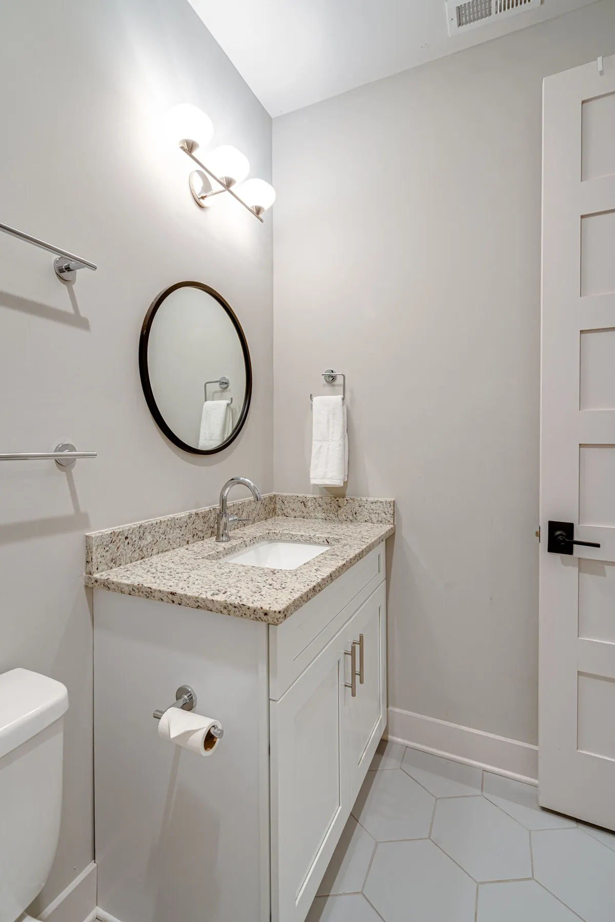 A fresh, minimalistic bathroom with a granite countertop, round mirror, and modern light fixtures. The white cabinetry and clean lines evoke a sense of relaxation and practicality, suggesting a well-maintained environment for personal care.