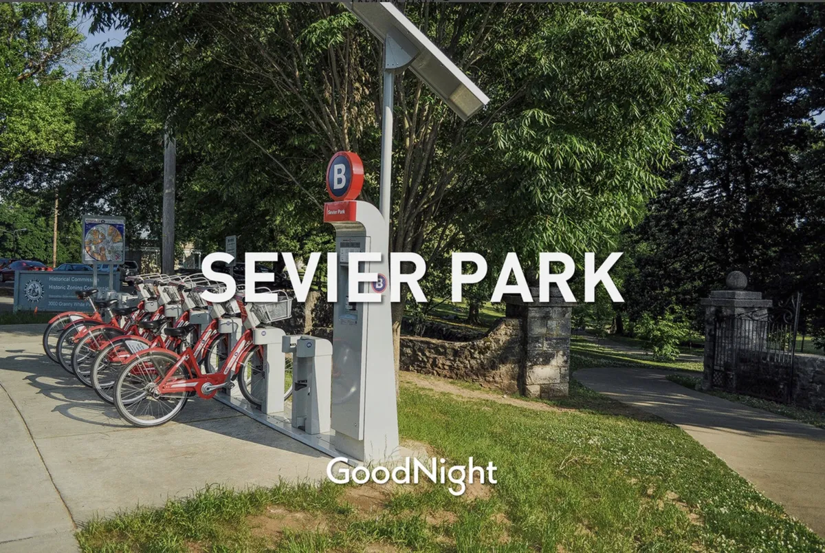 An inviting park entrance with a bike rental station, prominent park signage, and lush greenery. The area is set up for outdoor leisure and exploration, inviting visitors to stroll, bike, or picnic in the open space.